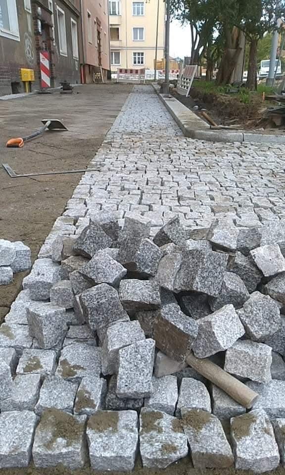 Ułożona część granitowej kostki brukowej na ścieżce, z narzędziami w tle. Część kostki jeszcze nieułożona znajduje się na pierwszym planie.