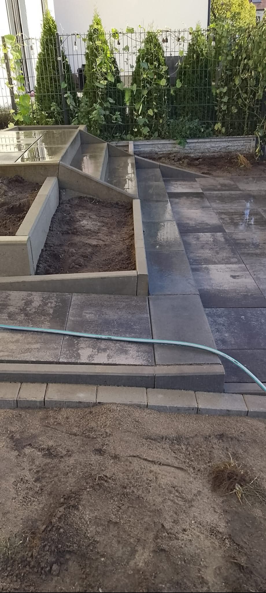 Ułożona kostka brukowa tworząca taras z podwyższonymi rabatami. Widoczne schody z kostki i elementy ogrodzenia. W tle zieleń roślin. Wąż ogrodowy leży na kostce.