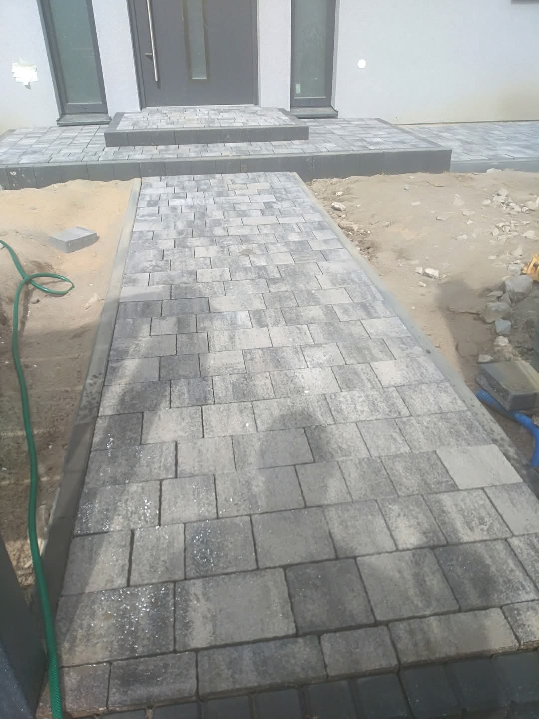 Ułożony chodnik z szarej kostki brukowej prowadzący do wejścia domu. Widoczne etapy prac ziemnych i wykończeniowych.
