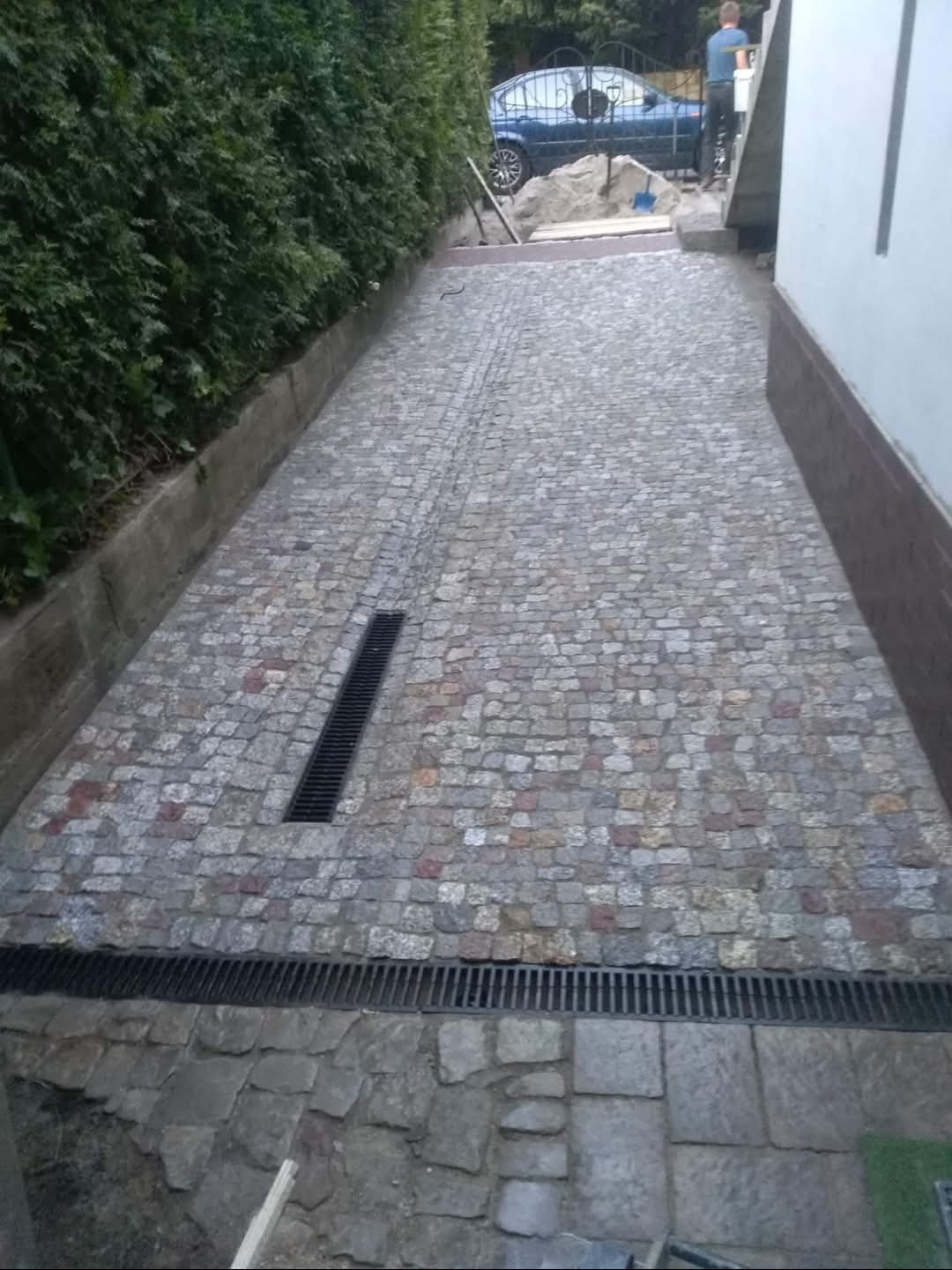 Ułożona kostka granitowa tworząca ścieżkę z odpływami liniowymi. W tle zieleń i częściowo widoczny samochód. Kąt ujęcia z dołu, podkreślający perspektywę.