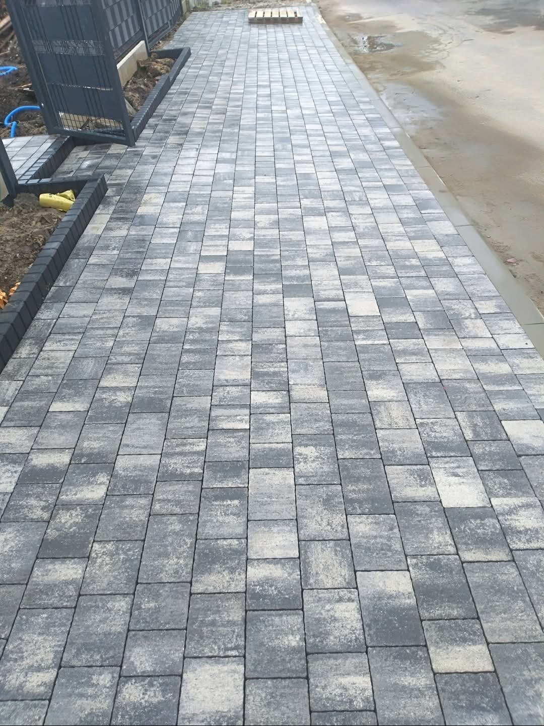 Ułożona szara kostka brukowa tworzy chodnik z obramowaniem. Widoczny fragment ogrodzenia i studzienka kanalizacyjna. Estetyczne wykończenie.
