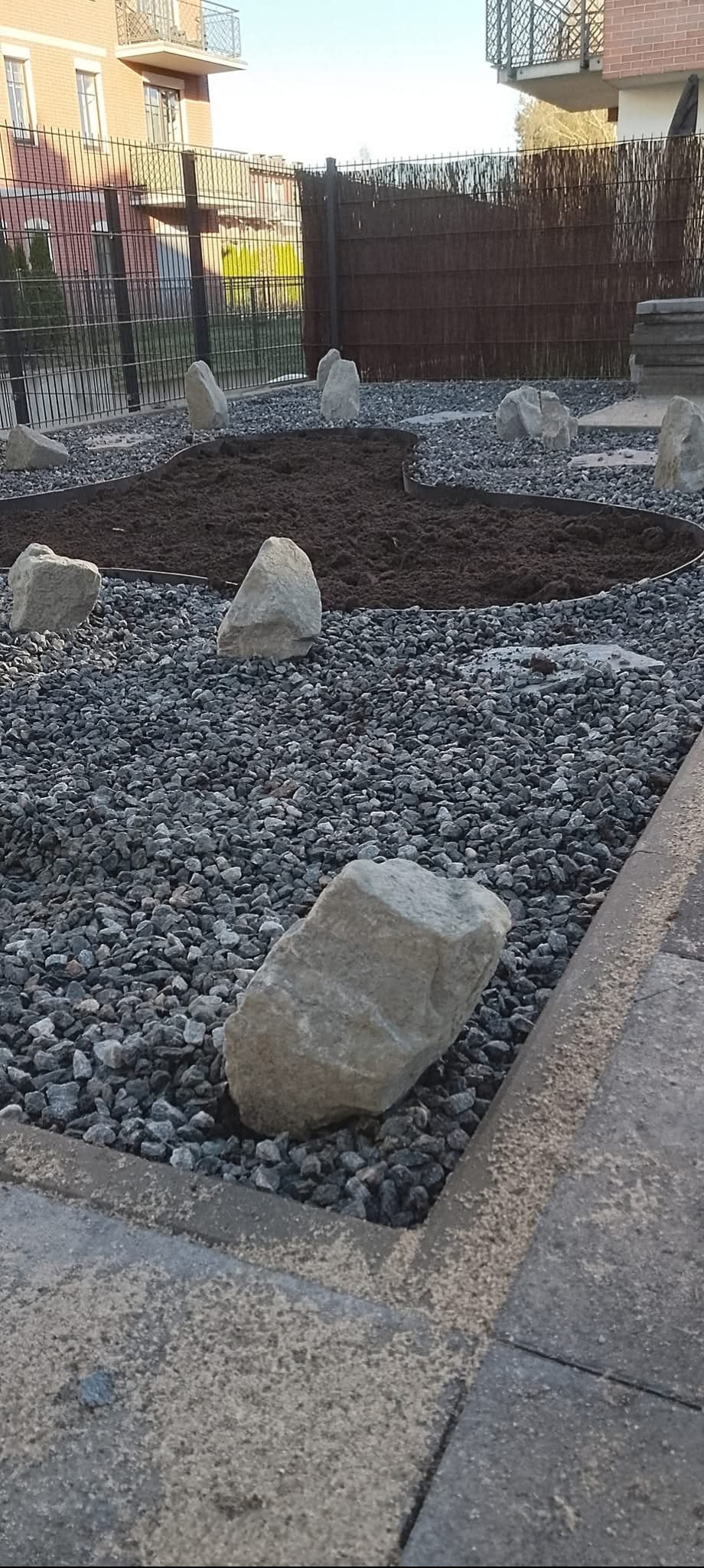 Ogród z szarym żwirem, ziemią i kamieniami. Metalowe obrzeże oddziela ziemię. W tle budynek z balkonami i ogrodzenie.