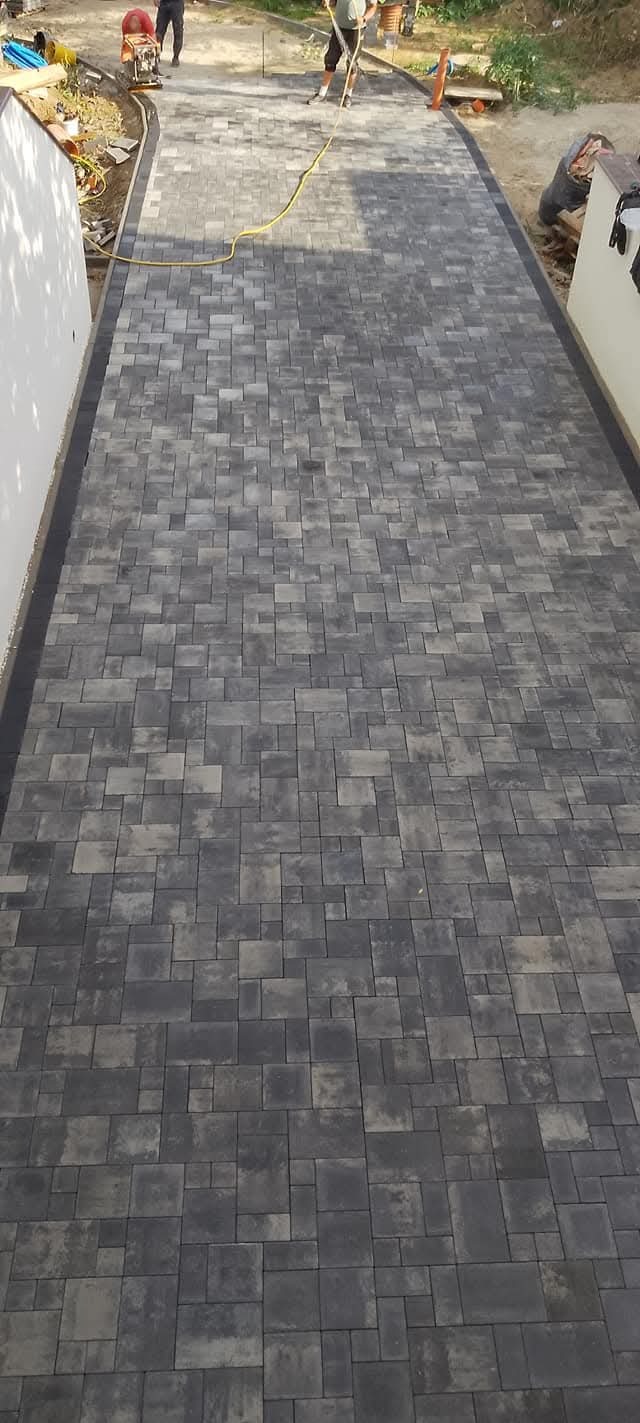 Ukończona ścieżka z szarej kostki brukowej ułożonej we wzór, widok z góry. W tle pracownicy i sprzęt budowlany.