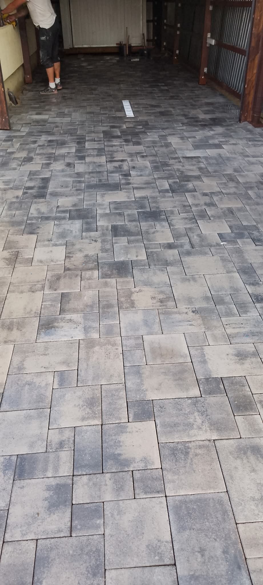 Ułożona szara kostka brukowa w nieregularny wzór, widok z góry. Ciemne fugi podkreślają kształt kostek. W tle brama garażowa i częściowo widoczna sylwetka osoby.