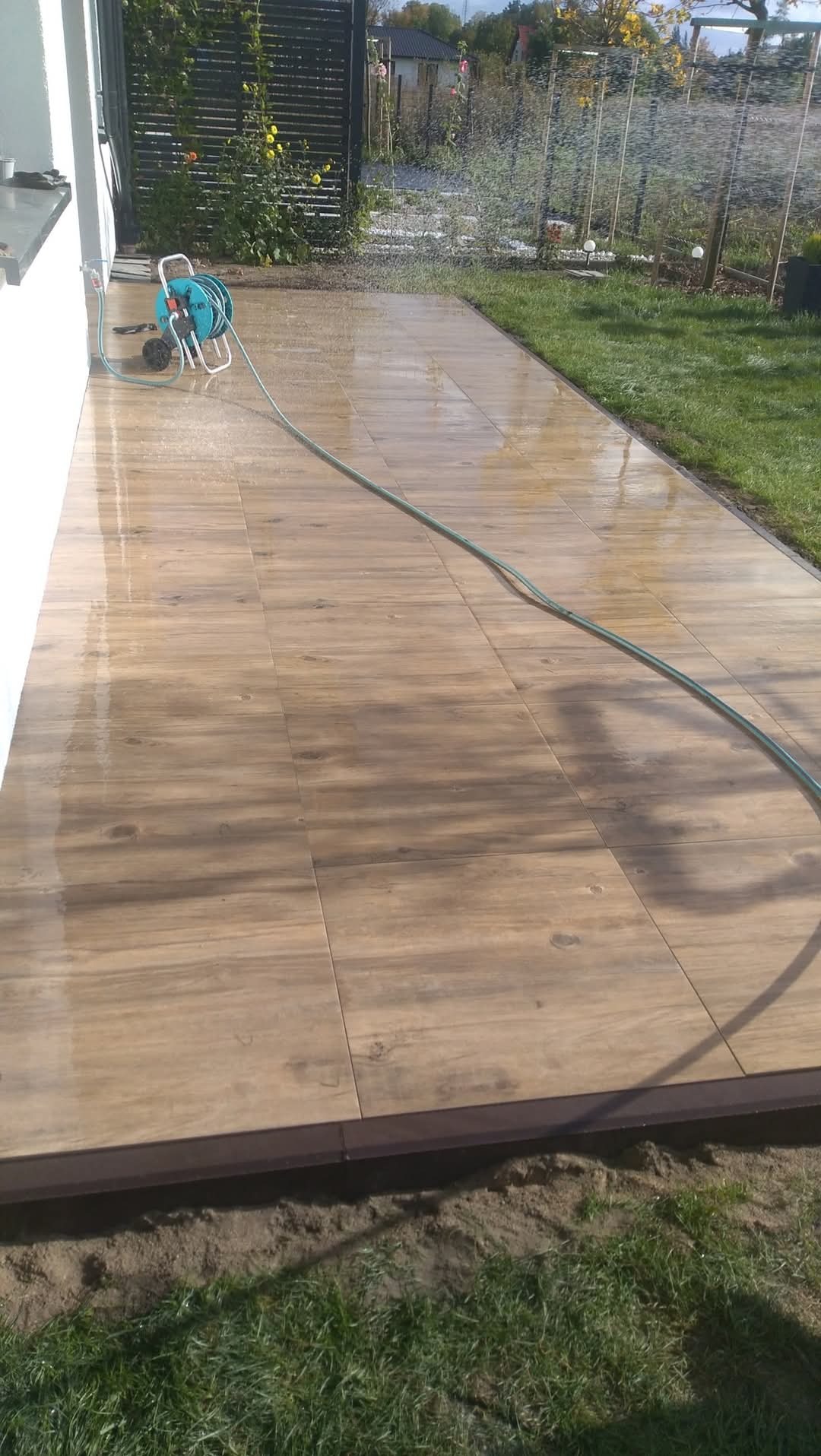 Umyta wodą z węża nowa, drewnopodobna, brązowa podłoga tarasu z delikatnym spadkiem, obok trawnik i ogrodzenie panelowe.