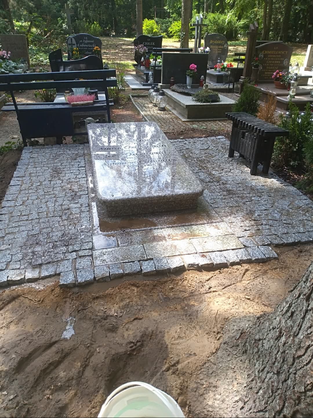 Granitowa kostka wokół nagrobka na cmentarzu, świeżo ułożona, z widocznymi śladami prac ziemnych i drzewem w tle. Kostka szara, nagrobek z polerowanego kamienia.