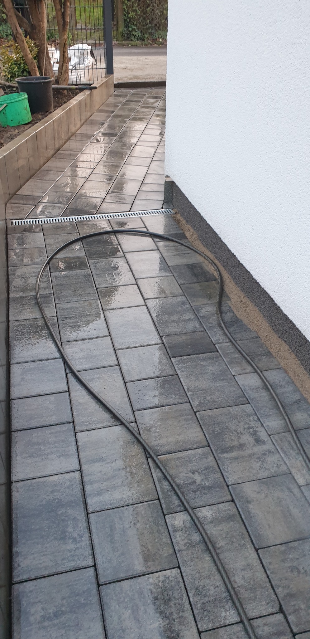 Ułożona, szara kostka brukowa z odpływem liniowym przy białej ścianie budynku. Na kostce leży zwinięty wąż. Widoczne donice i fragment ogrodzenia.