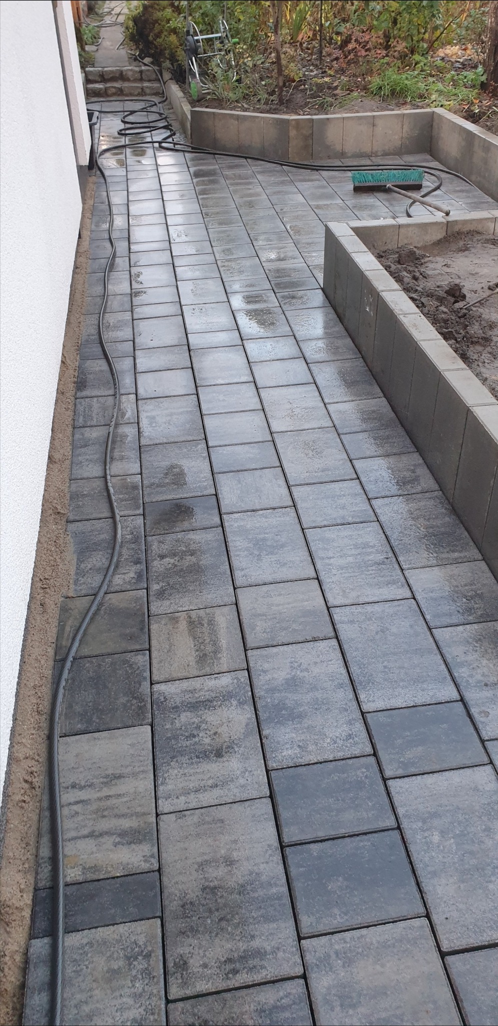 Świeżo ułożona, mokra kostka brukowa w odcieniach szarości tworzy ścieżkę przy budynku. Widoczny wąż ogrodowy i szczotka sugerują prace porządkowe po zakończeniu układania.