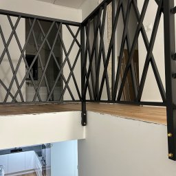 Rafał Kacica - Czarna, metalowa balustrada o geometrycznym wzorze krzyżowym, zamontowana na schodach w nowoczesnym wnętrzu. Widoczne detale mocowania i fragment kuchni w tle.