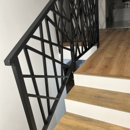 Rafał Kacica - Nowoczesna, czarna balustrada schodowa z geometrycznym wzorem, zamontowana na drewnianych schodach z białymi podstopnicami. Minimalistyczny design.