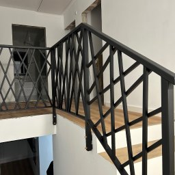 Rafał Kacica - Nowoczesna, czarna balustrada schodowa z geometrycznym wzorem rombów, zamontowana w jasnym wnętrzu. Widoczne drewniane stopnie i białe ściany.