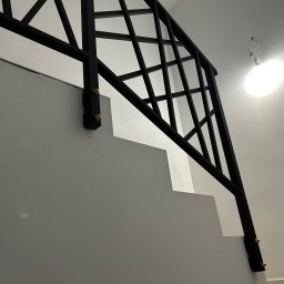 Rafał Kacica - Nowoczesna, czarna poręcz schodowa z geometrycznym wzorem, zamocowana do białych stopni. Widoczne złote śruby mocujące. Minimalistyczny design i oświetlenie.