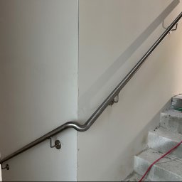 Rafał Kacica - Stalowa poręcz ze stali nierdzewnej, elegancko wygięta na narożniku ściany, zamontowana przy surowych betonowych schodach. Widoczne cienie i niedokończone ściany.