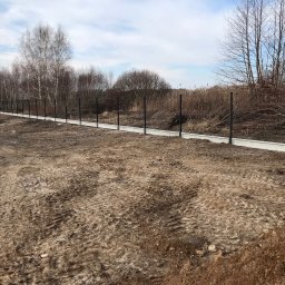 Mariusz Głąb - Długa linia betonowej podmurówki pod ogrodzenie z czarnymi słupkami, teren przygotowany pod dalszy montaż siatki, w tle drzewa bez liści i krzewy.