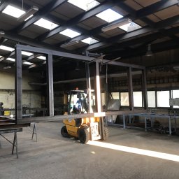 Mariusz Głąb - Wnętrze hali produkcyjnej: wózek widłowy transportuje metalową konstrukcję ramową, prawdopodobnie element przyszłego ogrodzenia, w tle widoczne stanowiska pracy i okna.