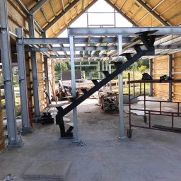 Mariusz Głąb - Wewnętrzna konstrukcja budynku w trakcie budowy z metalową konstrukcją schodów z czarnymi stopniami i srebrnymi wspornikami, widoczne metalowe słupy i drewniane panele ścian, w tle pojazd dostawczy.