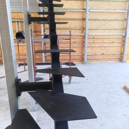 Mariusz Głąb - Konstrukcja stalowa schodów spiralnych w surowym stanie, przymocowana do metalowego słupa, widoczna na tle wznoszonej hali.