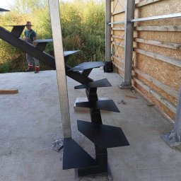 Mariusz Głąb - Surowe, metalowe schody zewnętrzne z kwadratowymi stopniami, w trakcie montażu na betonowej wylewce, widoczny pracownik w tle i konstrukcja szkieletowa budynku.