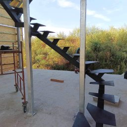 Mariusz Głąb - Konstrukcja schodów spiralnych z czarnego metalu w surowym stanie, widoczne rusztowanie i zieleń w tle, częściowo zmontowane.