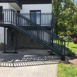 Mariusz Głąb - Metalowe schody zewnętrzne z balustradą, prowadzące na balkon budynku. Cień balustrady na kostce brukowej. Widok na zielony ogród w tle.