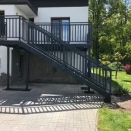 Metalowe schody zewnętrzne z balustradą, prowadzące na balkon budynku. Cień balustrady na kostce brukowej. Widok na zielony ogród w tle.