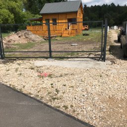 Mariusz Głąb - Nowoczesne ogrodzenie panelowe z bramą wjazdową, widok na drewniany dom i podwórko z podjazdem. Szara kolorystyka paneli i słupków, teren częściowo utwardzony.