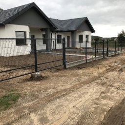 Mariusz Głąb - Częściowy widok montażu czarnego ogrodzenia panelowego wokół nowoczesnego domu z szarą dachówką, w pochmurny dzień. Widoczne betonowe podmurówki.