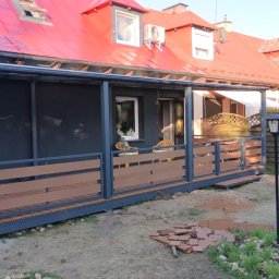 Mariusz Głąb - Nowoczesna, szara konstrukcja tarasu z brązowymi panelami na tle domu z czerwonym dachem. Widoczne elementy montażowe i fragmenty paneli na ziemi.