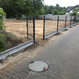Nowoczesne ogrodzenie panelowe z bramką, z betonową podmurówką, oddzielające posesję od chodnika z kostki brukowej. W tle dom jednorodzinny i zieleń.