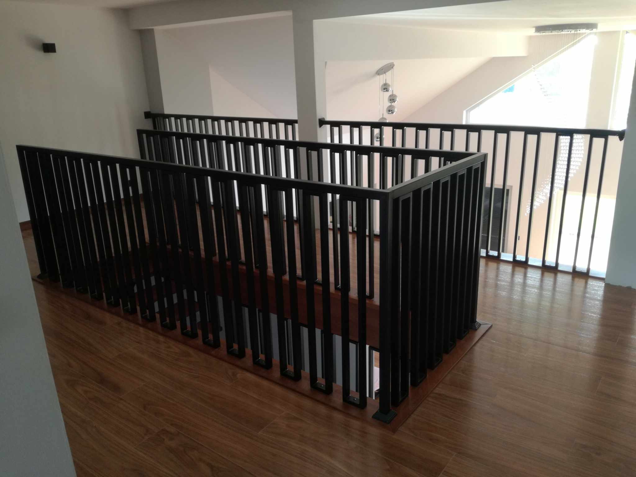 Nowoczesna balustrada schodowa z czarnych, pionowych prętów, zamontowana na drewnianych schodach w jasnym, przestronnym wnętrzu z widokiem na okno.