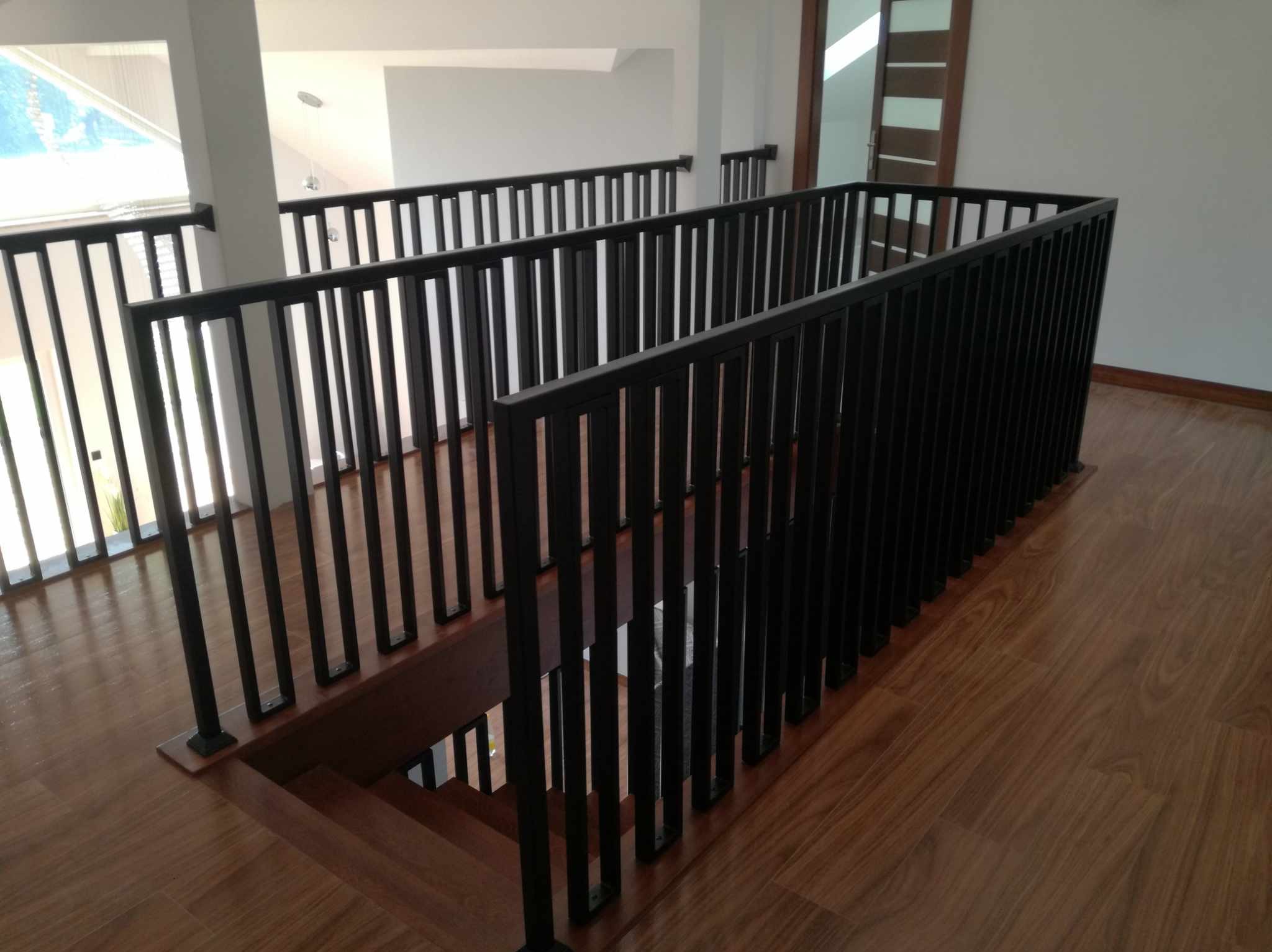 Nowoczesna, czarna balustrada schodowa z pionowych, prostokątnych profili, zamontowana wewnątrz budynku z drewnianymi schodami i podłogą.