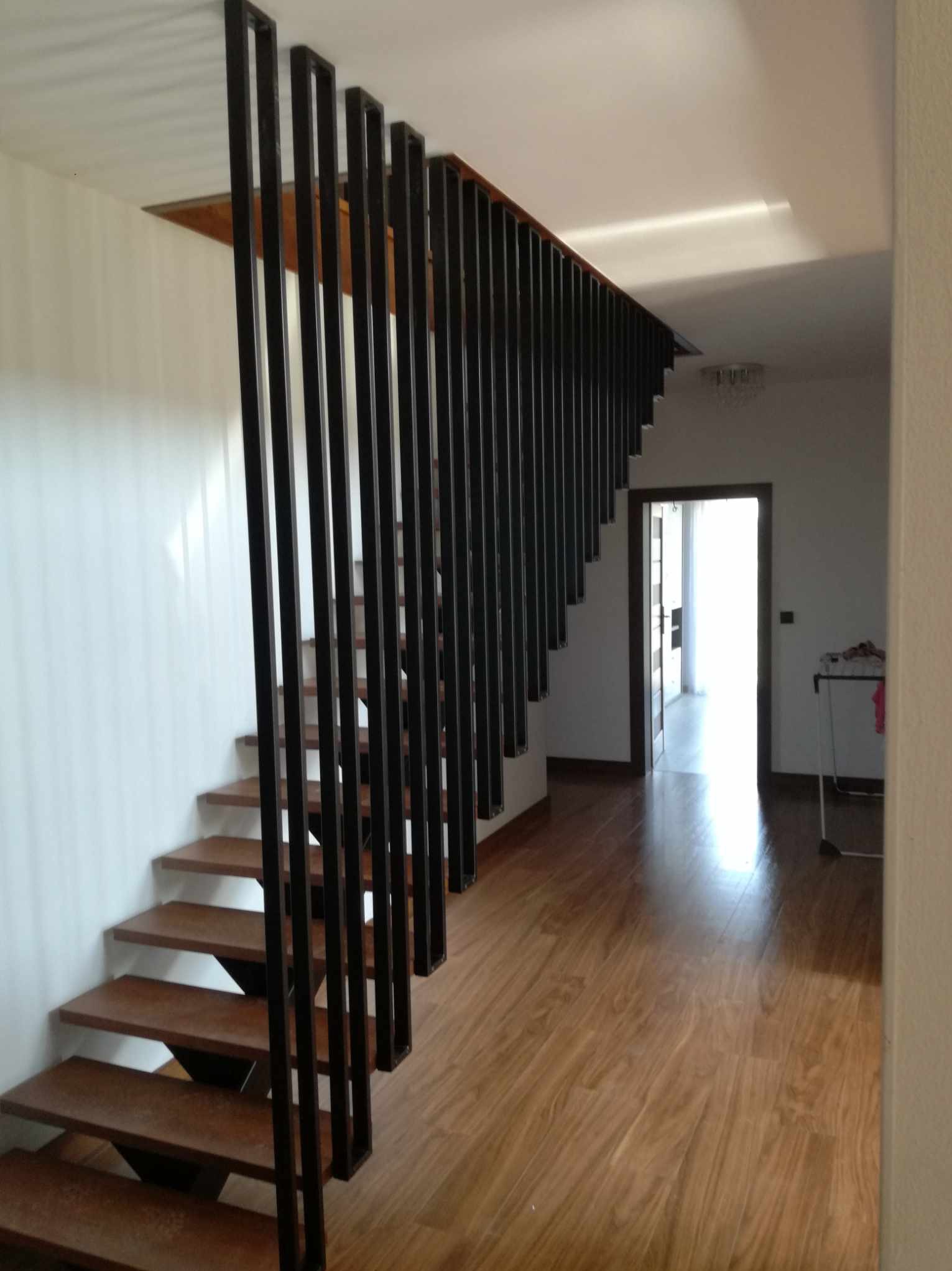 Schody drewniane z nowoczesną, metalową balustradą z pionowych, czarnych profili, widok z dołu na otwarte drzwi i dalszą część domu.