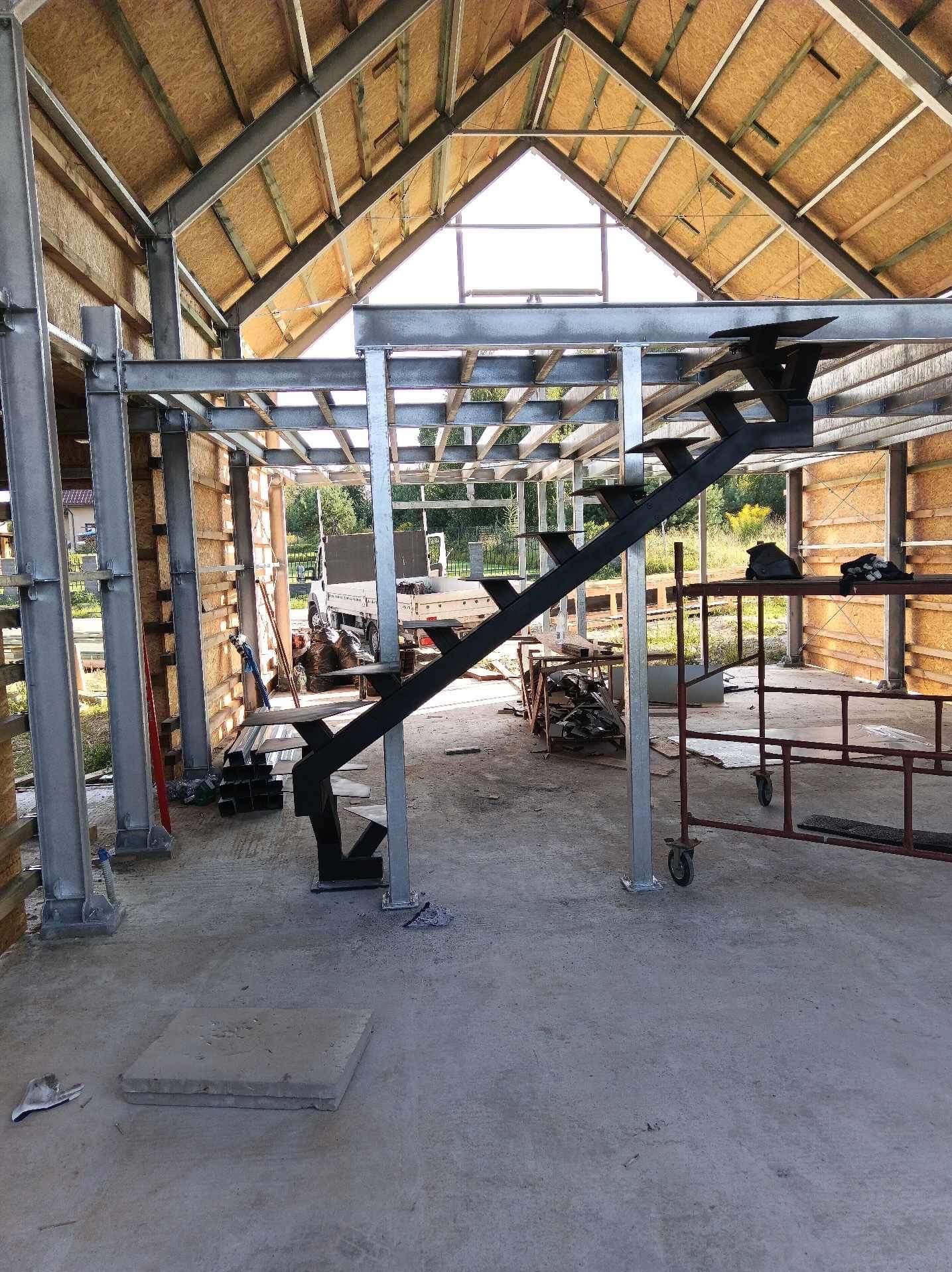 Wewnętrzna konstrukcja budynku w trakcie budowy z metalową konstrukcją schodów z czarnymi stopniami i srebrnymi wspornikami, widoczne metalowe słupy i drewniane panele ścian, w tle pojazd dostawczy.