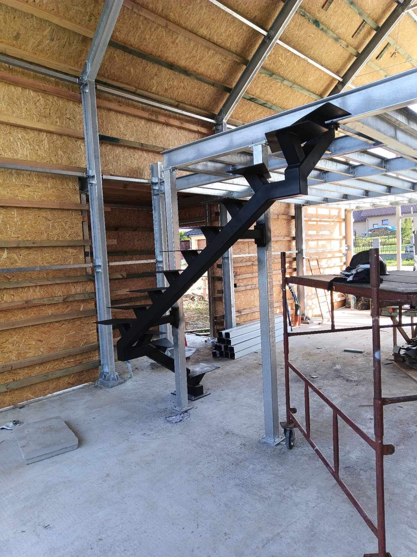 Stalowa konstrukcja schodów zewnętrznych w trakcie montażu, widoczne elementy konstrukcyjne budynku i rusztowanie.