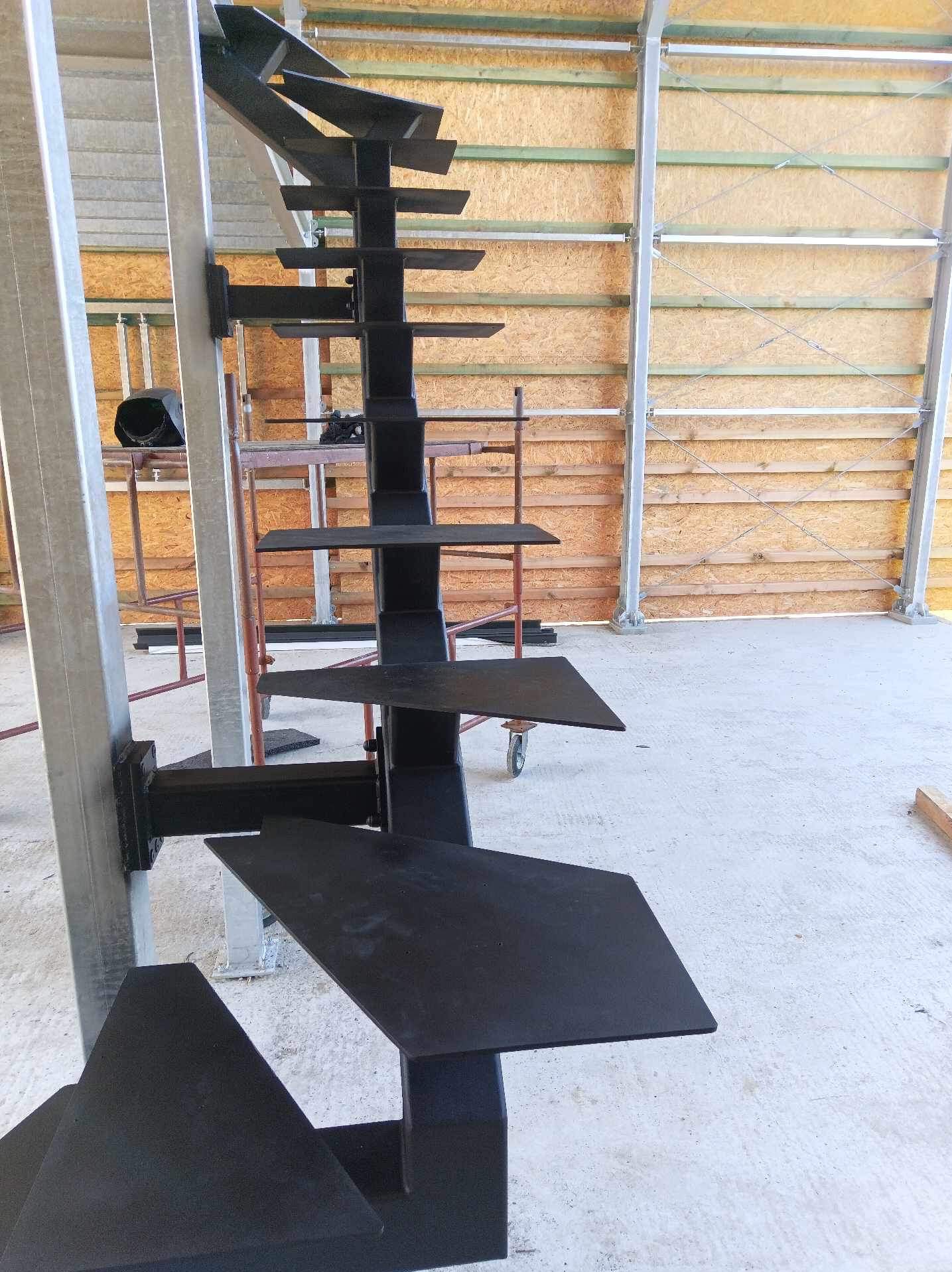 Konstrukcja stalowa schodów spiralnych w surowym stanie, przymocowana do metalowego słupa, widoczna na tle wznoszonej hali.