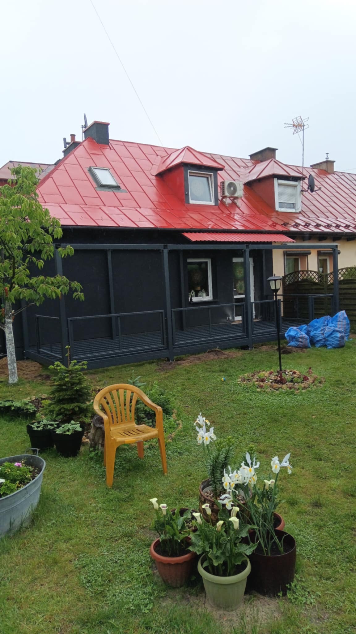 Nowoczesna, czarna konstrukcja ogrodowa z metalu, przylegająca do domu z czerwonym dachem. Uwagę zwraca żółte krzesło na tle zielonego trawnika.