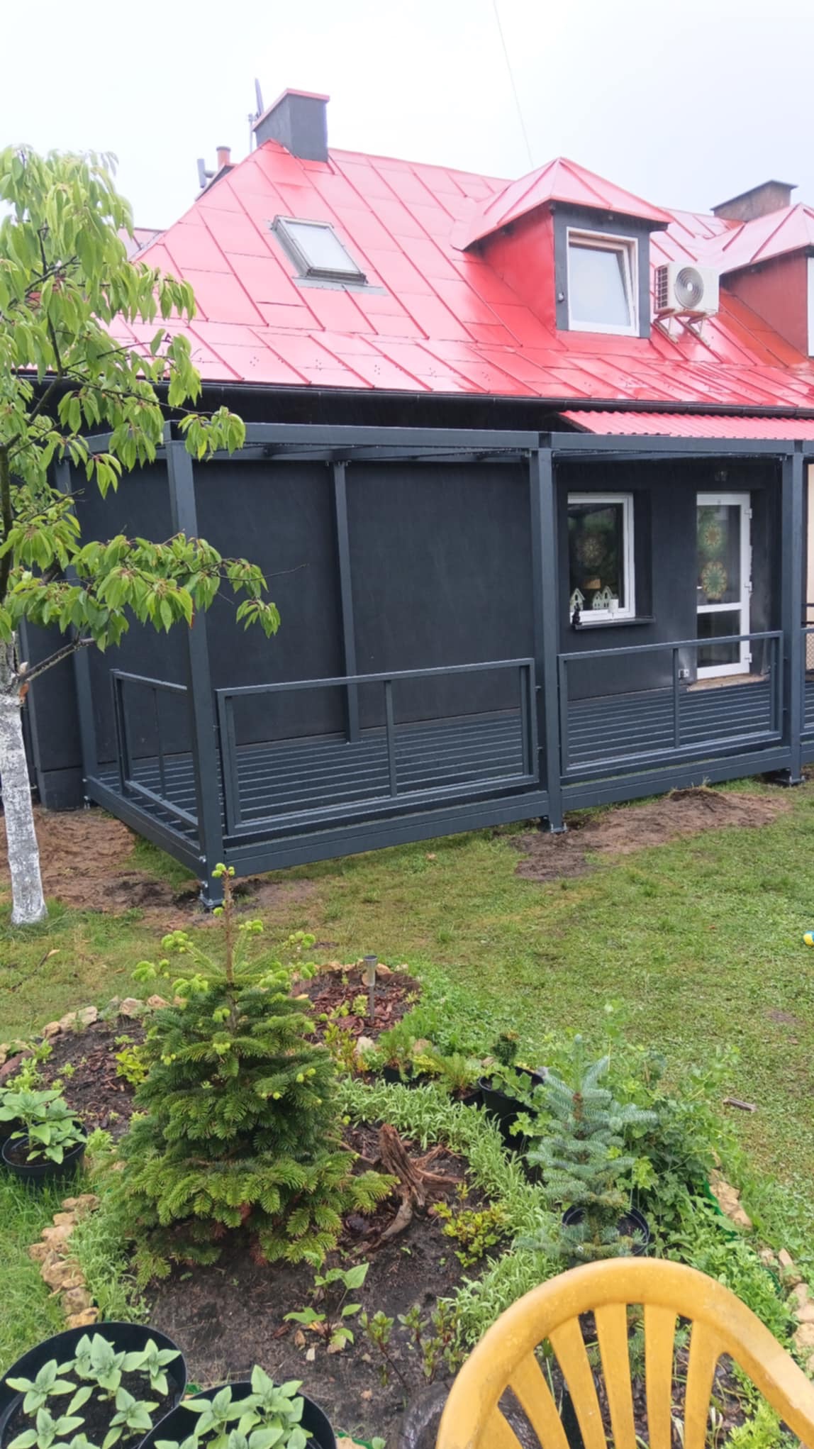 Nowoczesna, grafitowa pergola z metalowymi panelami, przylegająca do domu z czerwonym dachem. W tle zielony trawnik i rabata z iglakami.