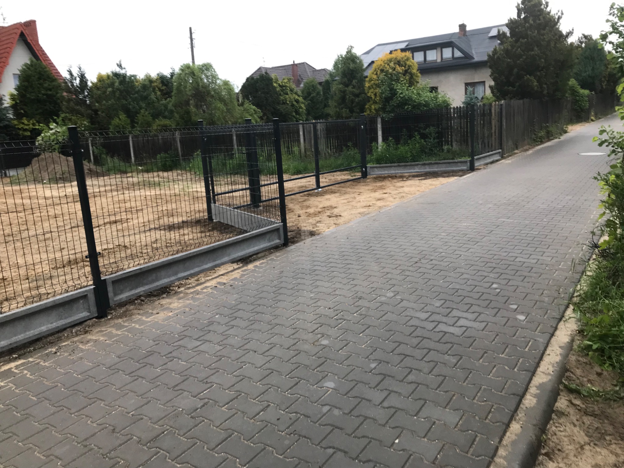 Nowoczesne ogrodzenie panelowe z betonową podmurówką i bramą wjazdową, widoczne z perspektywy chodnika. Dom i zieleń w tle.