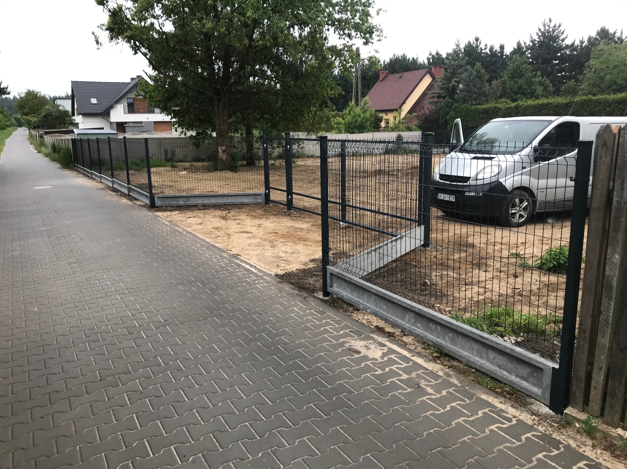 Nowoczesne, czarne ogrodzenie panelowe z podmurówką betonową, widok z zewnątrz posesji, szara kostka brukowa, srebrny van z otwartymi drzwiami w tle.