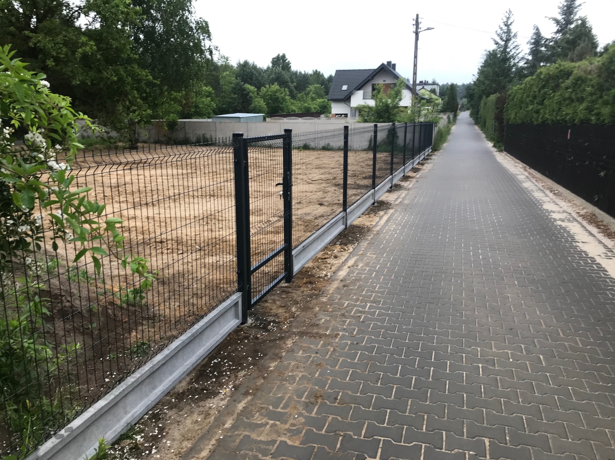 Nowoczesne ogrodzenie panelowe z podmurówką betonową, brama wjazdowa, widok z boku na tle posesji i drogi z kostki brukowej.