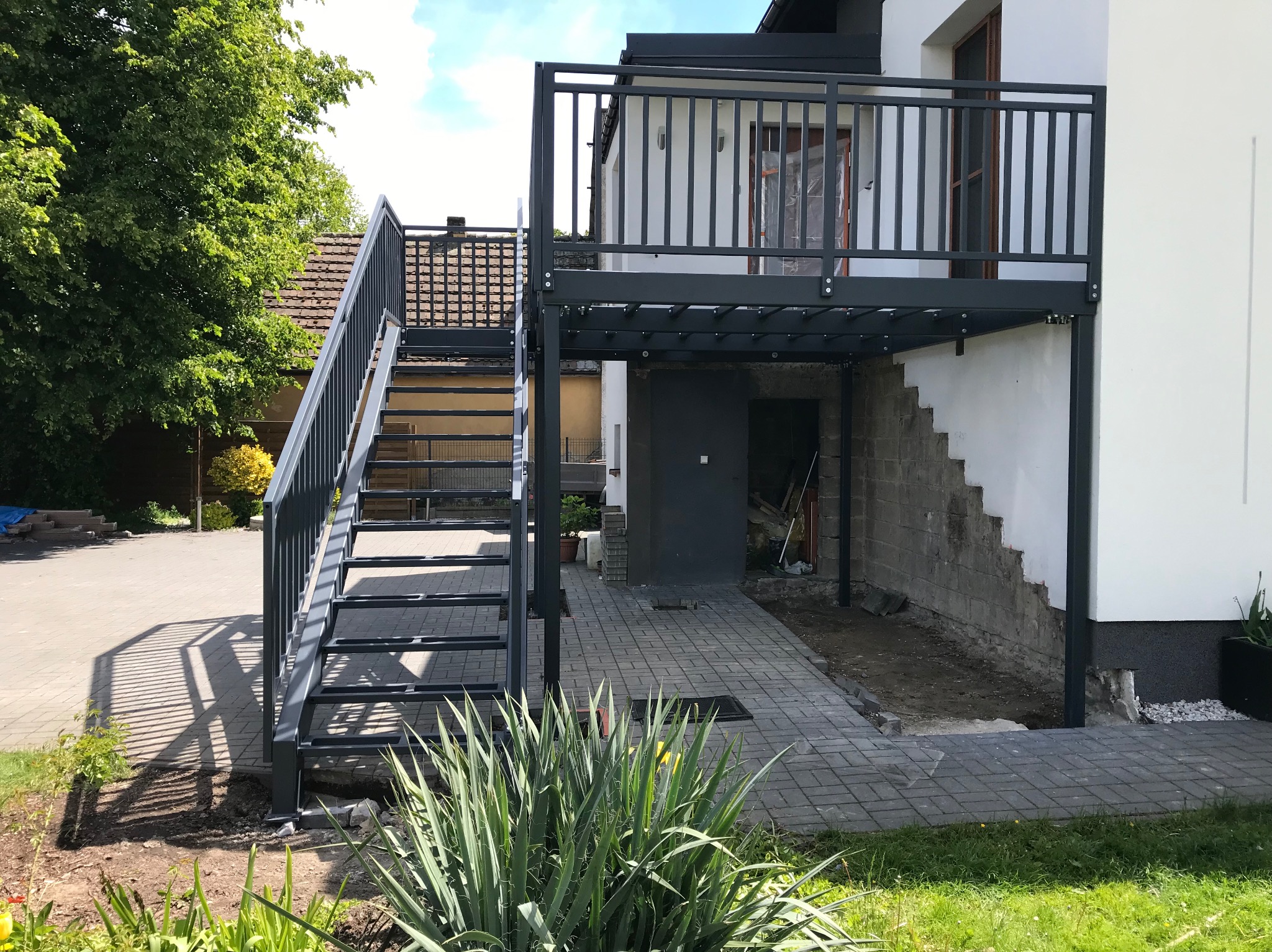 Nowoczesna konstrukcja metalowa z schodami i balkonem przy budynku. Szare elementy kontrastują z bielą elewacji. Widoczne detale spawania i montażu.