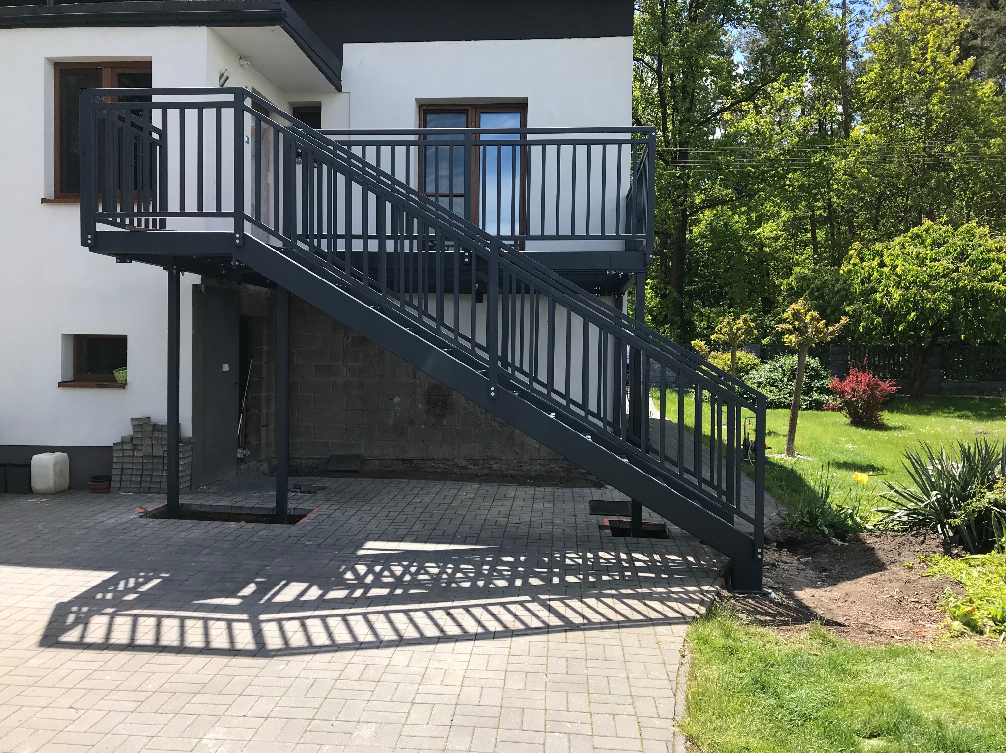 Metalowe schody zewnętrzne z balustradą, prowadzące na balkon budynku. Cień balustrady na kostce brukowej. Widok na zielony ogród w tle.