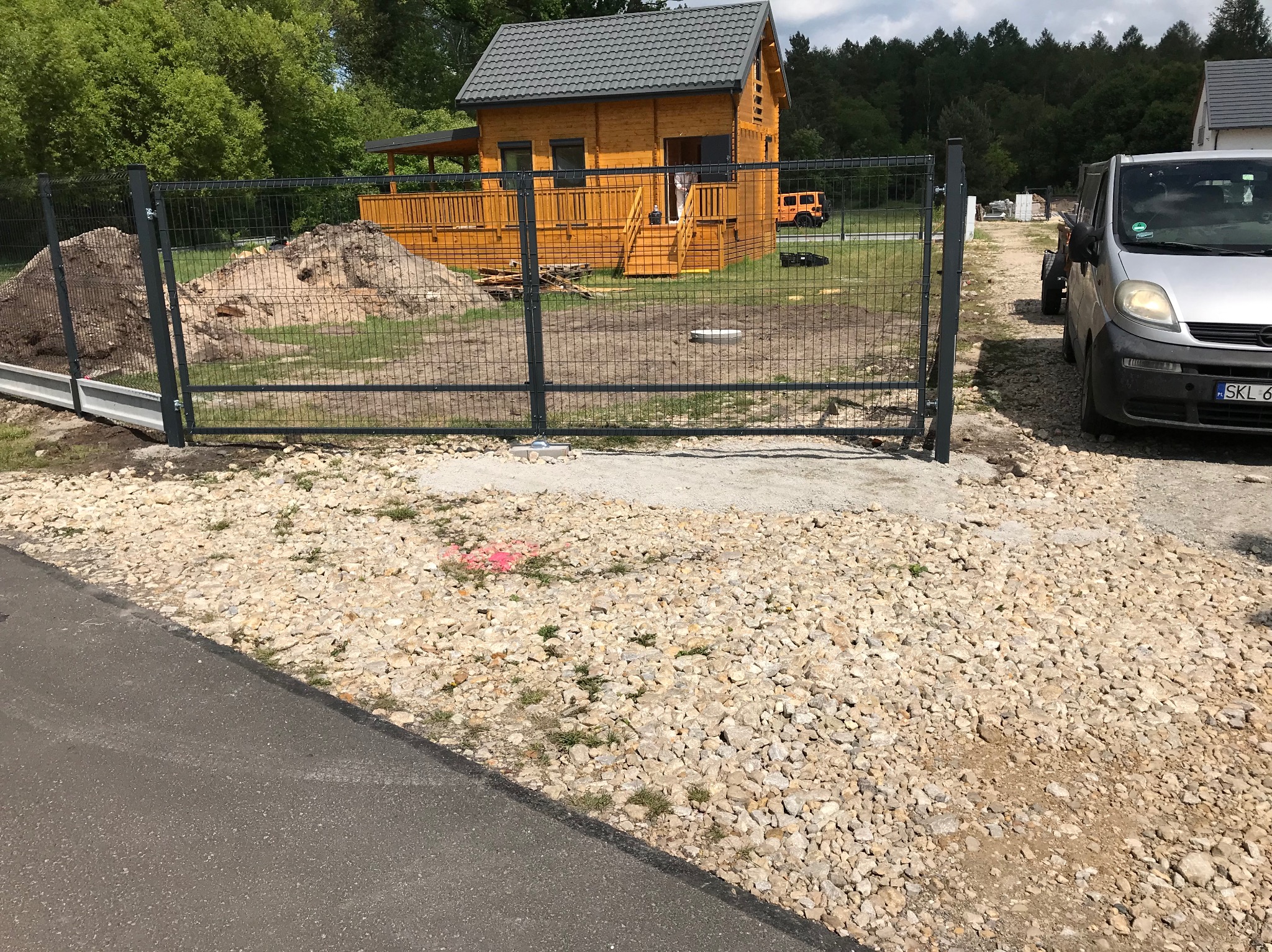 Nowoczesne ogrodzenie panelowe z bramą wjazdową, widok na drewniany dom i podwórko z podjazdem. Szara kolorystyka paneli i słupków, teren częściowo utwardzony.