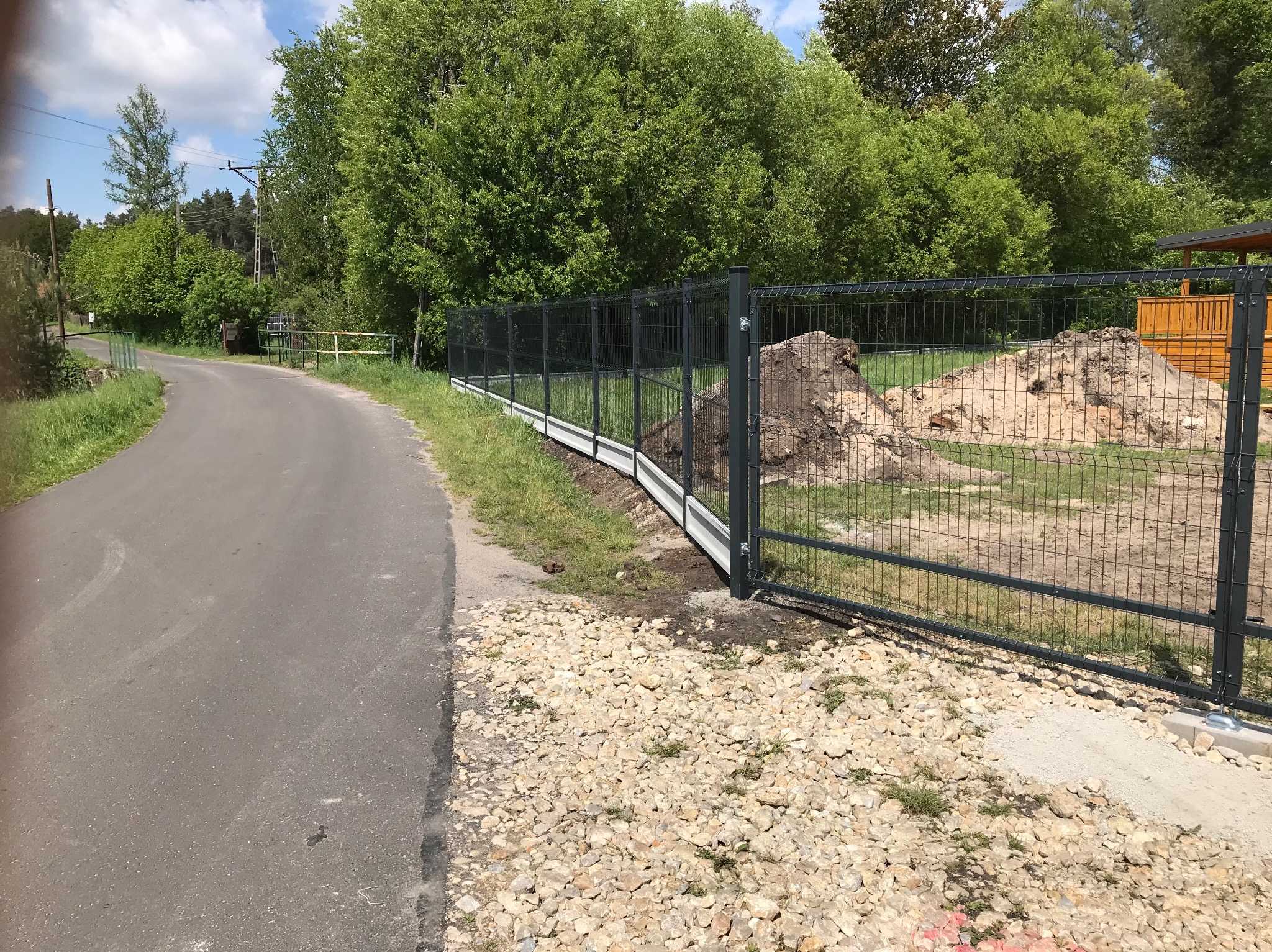 Nowoczesne, grafitowe ogrodzenie panelowe z podmurówką betonową, widoczne od strony drogi, z nasypem ziemi za nim i zielenią w tle. Ujęcie z poziomu gruntu.