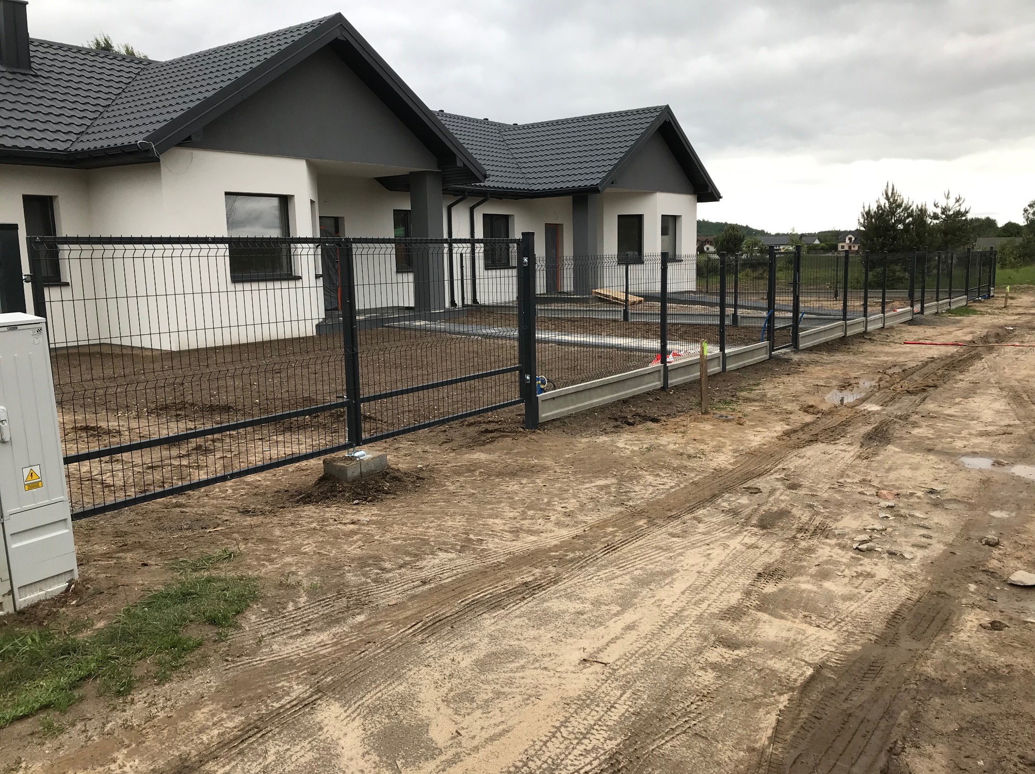 Częściowy widok montażu czarnego ogrodzenia panelowego wokół nowoczesnego domu z szarą dachówką, w pochmurny dzień. Widoczne betonowe podmurówki.