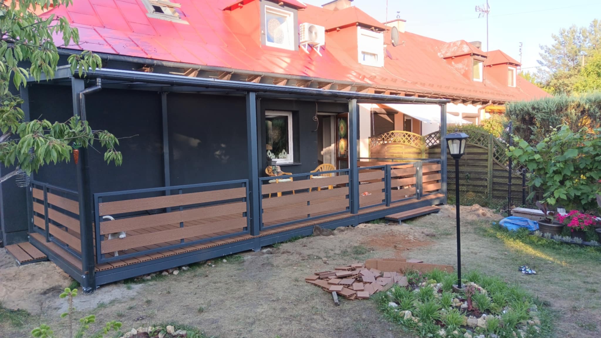 Zadaszony taras z brązowymi panelami i szarą ramą, przylegający do ciemnego domu z czerwonym dachem. Widoczne elementy wykończeniowe i lampa solarna w ogrodzie.