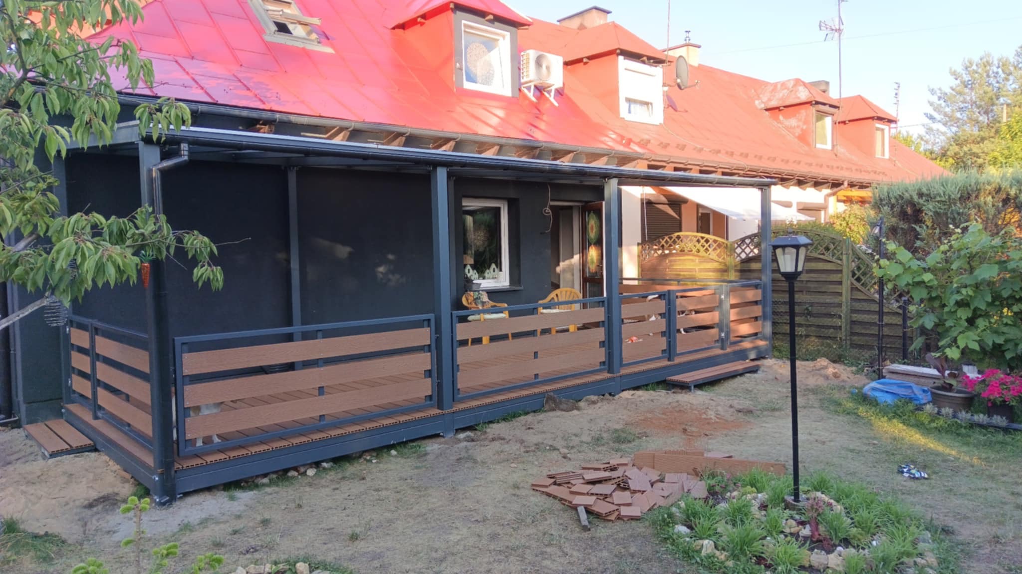 Nowoczesna, szara konstrukcja tarasu z brązowymi panelami na tle domu z czerwonym dachem. Widoczne elementy montażowe i fragmenty paneli na ziemi.