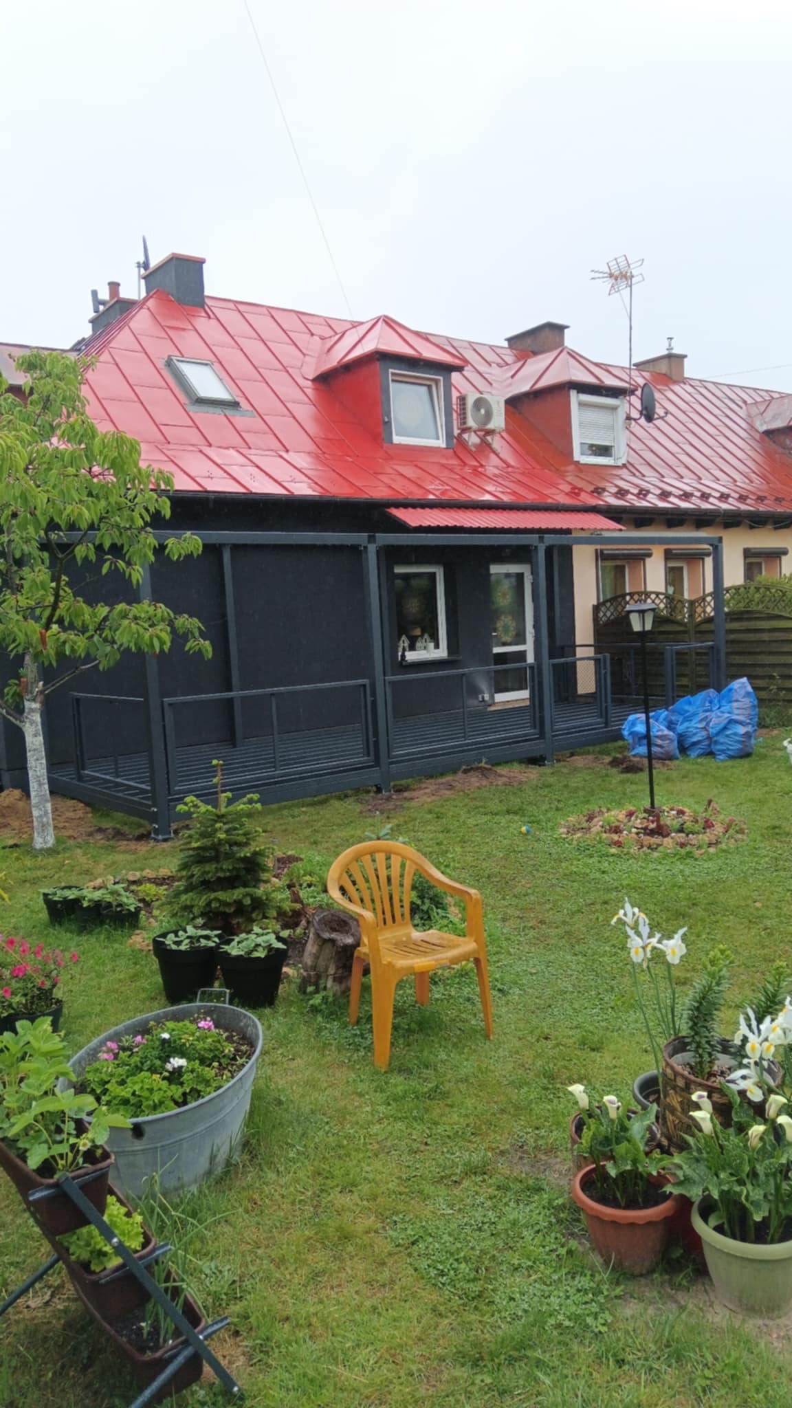 Nowoczesna, grafitowa pergola z metalową konstrukcją i drewnianym podestem, przylegająca do domu z czerwoną dachówką. Ustawiona w ogrodzie z żółtym krzesłem i kwiatami.