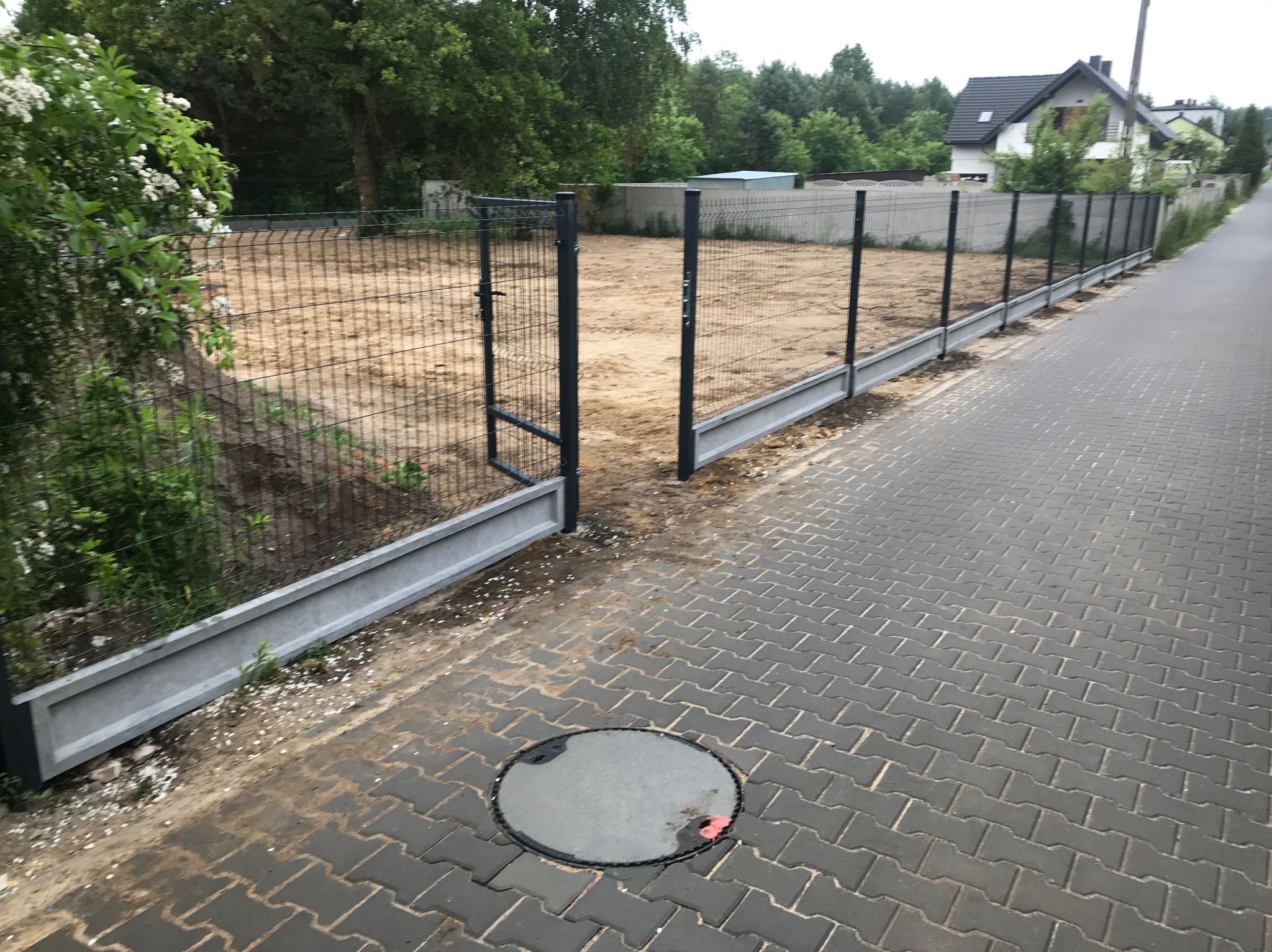 Nowoczesne ogrodzenie panelowe z bramką, z betonową podmurówką, oddzielające posesję od chodnika z kostki brukowej. W tle dom jednorodzinny i zieleń.