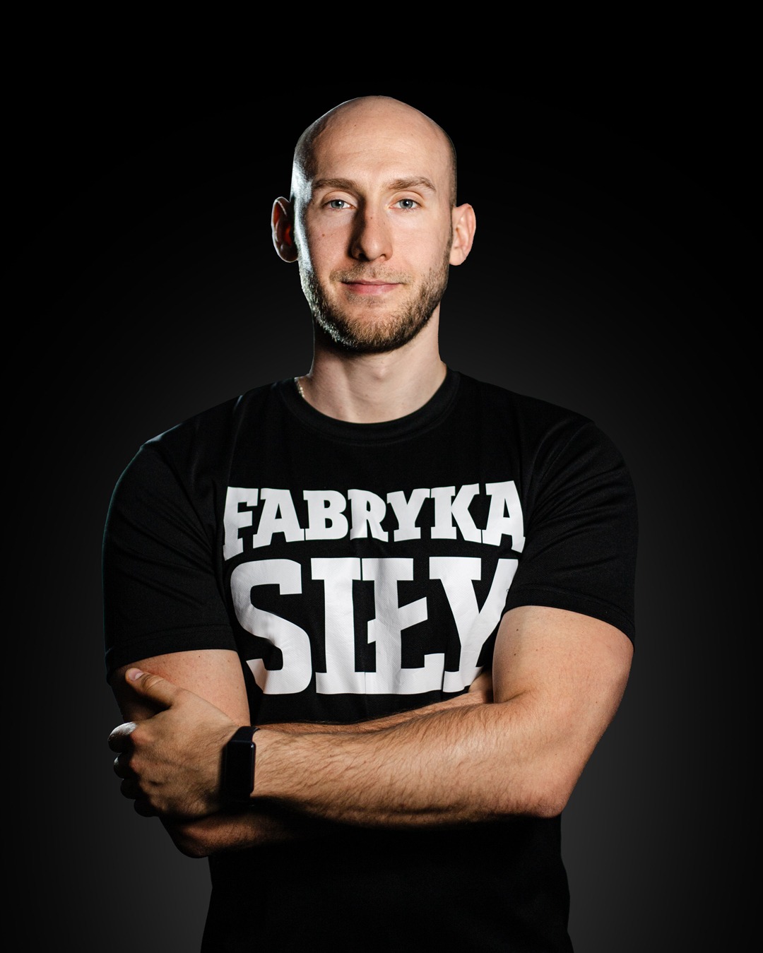 Portret mężczyzny z brodą w czarnej koszulce z napisem 'Fabryka Siły', skrzyżowane ramiona, smartwatch na nadgarstku, ciemne tło.